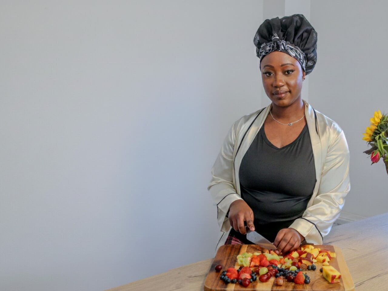Woman preparing a fruit salad on a wooden cutting board with a neutral background wearing a lew's collections hair bonnet with the marble noir design