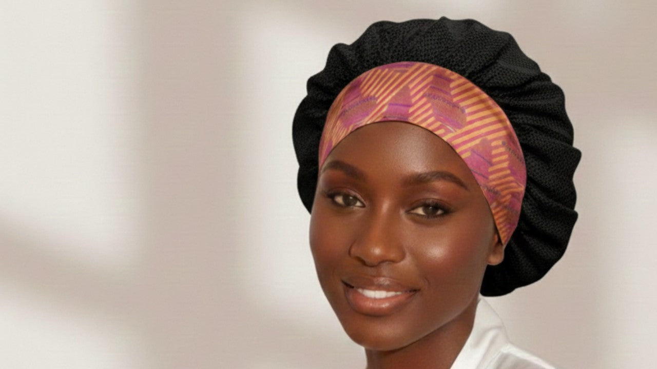 Black woman wearing a black satin hair bonnet with a Aztec Afrique pattern and standard band, set against a light background with subtle shadow rays